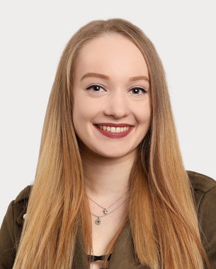 Eine junge Frau mit langen, blonden Haaren und einem freundlichen Lächeln trägt eine grüne Jacke. Der Hintergrund ist neutral und hell.