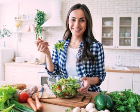 Eine Frau in einer karierten Bluse hält eine Schüssel mit Salat und lächelt. Auf der Küchentheke sind frisches Gemüse und Kräuter sichtbar.