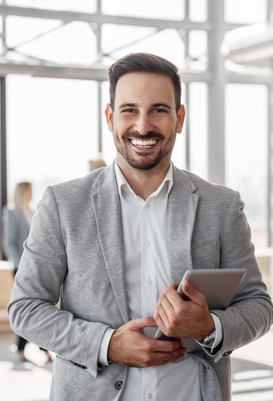 Ein lächelnder Mann in einem grauen Anzug hält ein Tablet in der Hand. Im Hintergrund sind mehrere Personen in einem modernen Büro zu sehen, die miteinander sprechen.