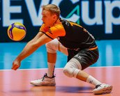 Ein Volleyballspieler in einem orange-schwarzen Trikot führt einen Pass aus, während er sich auf dem Spielfeld befindet. Im Hintergrund ist ein Banner mit der Aufschrift "CEV Cup" zu sehen.