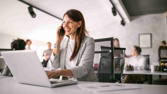Eine Frau spricht am Telefon, während sie an einem Laptop arbeitet. Im Hintergrund sind weitere Personen in einem modernen Büro zu sehen.