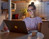 Eine junge Frau mit Brille sitzt an einem Tisch, nutzt einen Laptop und hört mit Kopfhörern Musik oder einen Podcast.