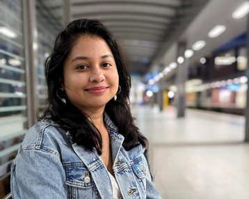 Eine junge Frau mit langen, dunklen Haaren sitzt auf einer Bank, trägt eine Jeansjacke und lächelt in einem modernen Innenraum.
