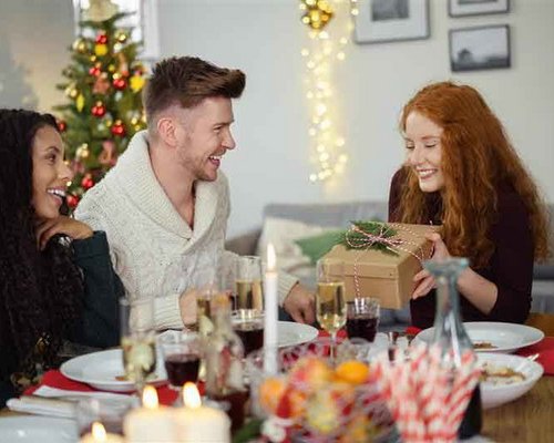 Eine Gruppe von vier Personen sitzt an einem festlich gedeckten Tisch, während eine Frau mit roten Haaren ein Geschenk überreicht. Im Hintergrund ist ein Weihnachtsbaum und festliche Dekoration sichtbar.