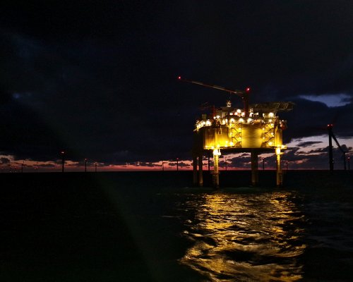 Eine Ölplattform leuchtet in der Dämmerung, umgeben von Wasser und dunklen Wolken. Der Himmel zeigt einen Farbverlauf von Dunkelblau zu Orange am Horizont.