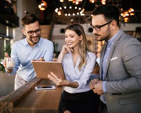 Drei Personen in einem modernen Café betrachten gemeinsam ein Tablet. Zwei Männer, einer mit Brille, halten eine Tasse, während die Frau lächelt und auf den Bildschirm zeigt.