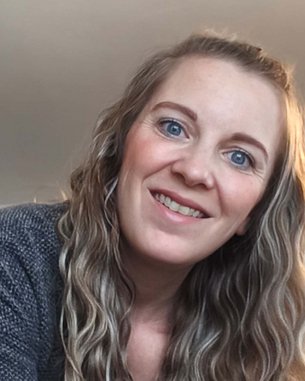 Eine Frau mit langen, lockigen Haaren lächelt in die Kamera, während sie sich leicht nach vorne beugt. Sie trägt einen grauen Pullover.