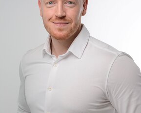 Ein Mann mit rotem Haar und Bart trägt ein weißes Hemd und lächelt in die Kamera. Der Hintergrund ist hell und neutral.