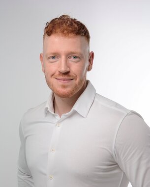 Ein Mann mit rotem Haar und Bart trägt ein weißes Hemd und lächelt in die Kamera. Der Hintergrund ist hell und neutral.