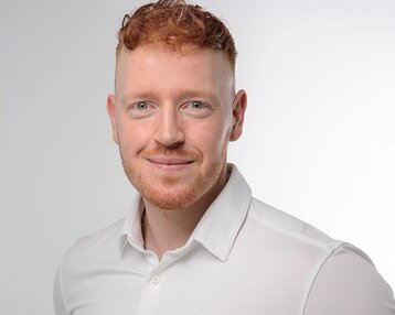 Ein Mann mit rotem Haar und Bart trägt ein weißes Hemd und lächelt in die Kamera. Der Hintergrund ist hell und neutral.