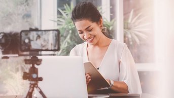 Eine Frau sitzt an einem Tisch, lächelt und hält ein Notizbuch in der Hand, während sie auf einen Laptop schaut. Ein Smartphone ist auf einem Stativ positioniert, um sie zu filmen.