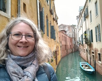 Eine Person mit lockigem, grauem Haar und einer Brille lächelt vor einem venezianischen Kanal, umgeben von bunten Gebäuden. Ein kleines Boot schwimmt im Wasser.
