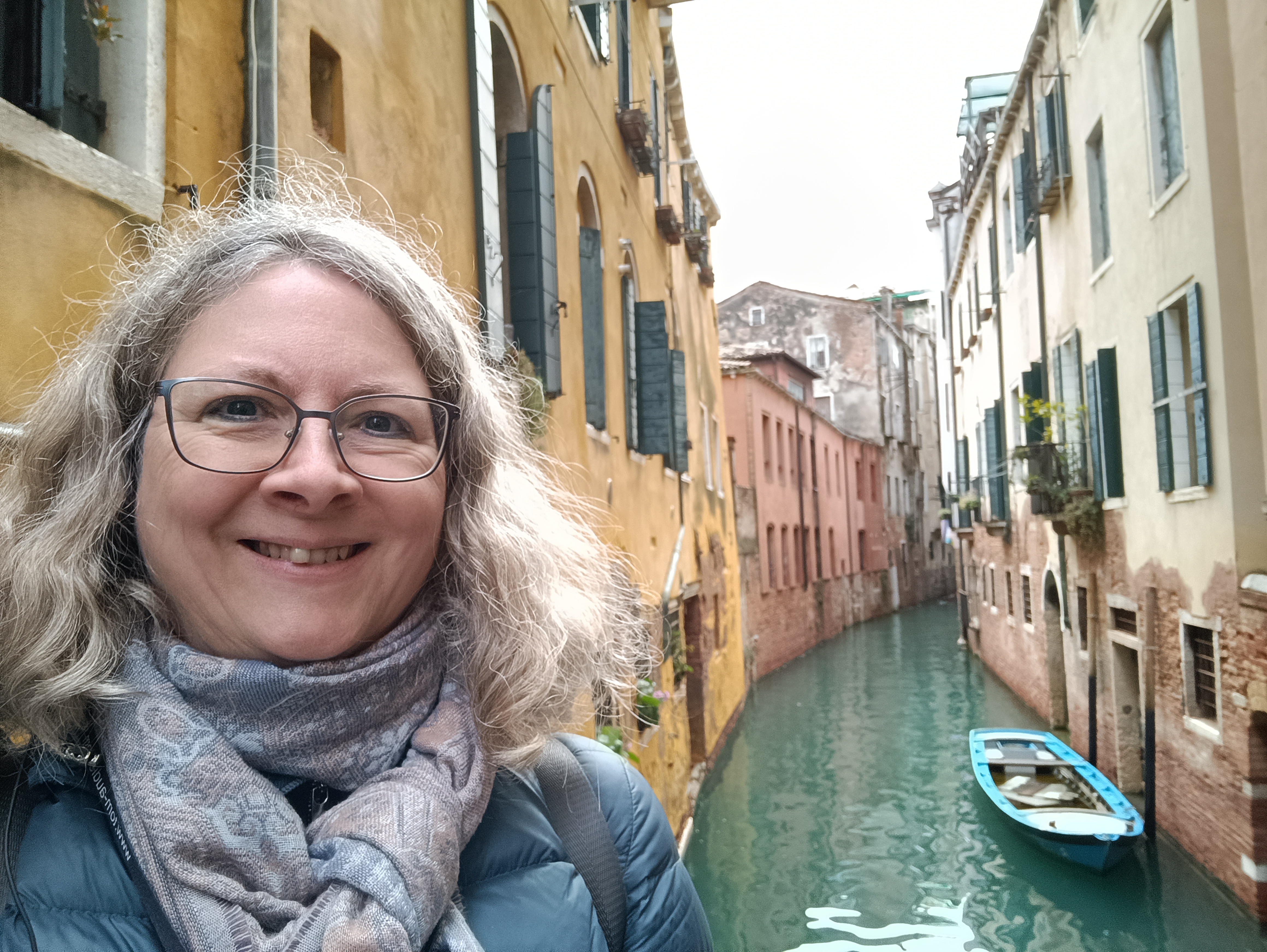 Eine Person mit lockigem, grauem Haar und einer Brille lächelt vor einem venezianischen Kanal, umgeben von bunten Gebäuden. Ein kleines Boot schwimmt im Wasser.