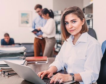 Eine Frau mit kurzen Haaren sitzt an einem Schreibtisch und arbeitet an einem Laptop. Im Hintergrund sind zwei Personen zu sehen, die miteinander sprechen, während ein Mann auf einem Stuhl sitzt und liest.