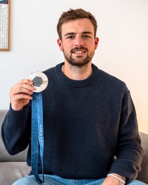 Ein Mann sitzt auf einem Sofa und hält eine Medaille mit blauer Schleife in der Hand. Im Hintergrund ist ein Kunstwerk an der Wand zu sehen.