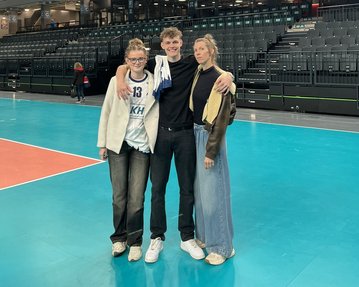 Drei Personen stehen auf einem Volleyballfeld in einer Halle. Eine Person trägt ein Trikot, während die anderen lässige Kleidung tragen.
