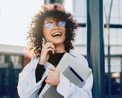 Eine lächelnde Frau mit lockigem Haar trägt eine weiße Bluse und eine Laptop-Tasche. Sie hält ein Tablet in der Hand und telefoniert, während sie an einem Pfosten steht.