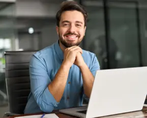 Ein lächelnder Mann sitzt an einem Schreibtisch vor einem Laptop in einem modernen Büro.
