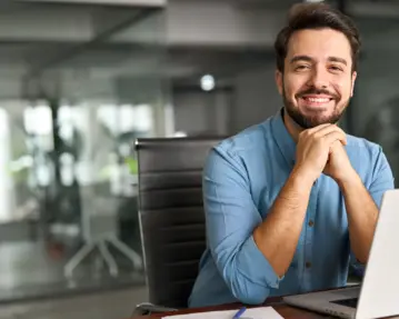 Ein lächelnder Mann sitzt an einem Schreibtisch vor einem Laptop in einem modernen Büro.