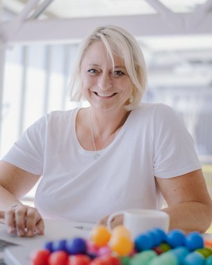 Marion Krüger-Altena, Studentin Psychologie im Online-Abendstudium (B.Sc.) | SRH Fernhochschule Eine lächelnde Frau mit blonden Haaren sitzt an einem Tisch mit einem Laptop und einer Tasse. Vor ihr liegt eine bunte Ansammlung von kleinen Kugeln.