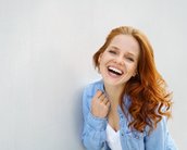 Eine lächelnde Frau mit langen, lockigen roten Haaren trägt ein hellblaues Hemd und steht vor einer neutralen Wand.