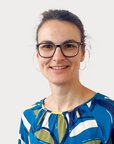 Eine Frau mit Brille trägt ein buntes, gemustertes Oberteil. Sie lächelt und steht vor einem neutralen Hintergrund.