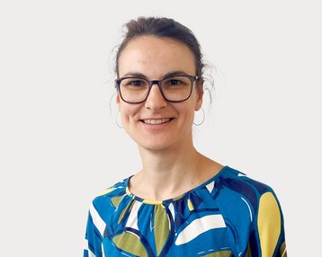 Eine Frau mit Brille trägt ein buntes, gemustertes Oberteil. Sie lächelt und steht vor einem neutralen Hintergrund.