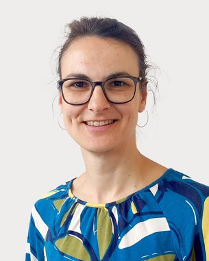 Eine Frau mit Brille trägt ein buntes, gemustertes Oberteil. Sie lächelt und steht vor einem neutralen Hintergrund.