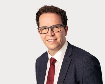 Ein Mann mit lockigem Haar trägt eine Brille und einen Anzug mit Krawatte. Er lächelt und steht vor einem neutralen Hintergrund.
