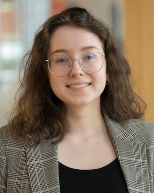 Junge Frau mit lockigem Haar und Brille, die ein kariertes Blazer-Jackett trägt. Sie steht in einem modernen Innenraum mit Holz- und Glaselementen.