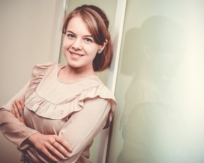 Eine junge Frau mit einem lächelnden Gesicht steht mit verschränkten Armen vor einer Wand. Sie trägt eine blusige, rüschige Bluse und hat eine elegante Frisur. Ihr Schatten ist leicht im Hintergrund sichtbar.