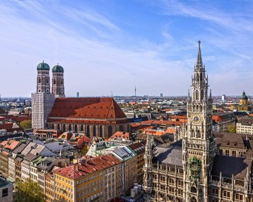 Moderne Gebäude mit großen Fenstern und einem überdachten Außenbereich. Tische und Stühle unter grünen Sonnenschirmen sind im Vordergrund sichtbar.