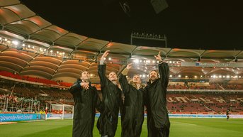Vier Personen in schwarzen Roben posieren im Stadion, während sie mit ihren Handys Selfies machen. Im Hintergrund sind die Tribünen und das Spielfeld sichtbar.