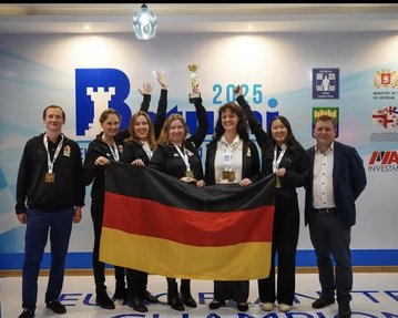 Ein Team von sieben Personen hält eine deutsche Flagge und posiert mit einem Pokal. Im Hintergrund sind Flaggen und Sponsorenlogos sichtbar.