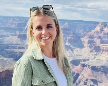 Eine Person mit langen, blonden Haaren und Sonnenbrille steht am Grand Canyon, mit einer beeindruckenden Landschaft im Hintergrund.