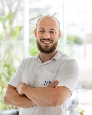 Ein Mann mit Bart und kurzen Haaren steht mit verschränkten Armen in einem hellen Raum. Er trägt ein weißes Hemd mit einem Logo auf der Brust und lächelt freundlich.