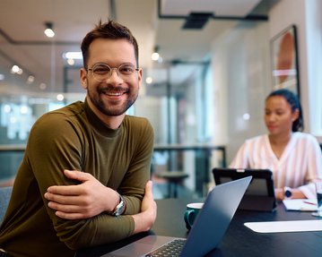 Ein lächelnder Mann mit Brille sitzt an einem Tisch mit einem Laptop, während eine Frau im Hintergrund an einem Tablet arbeitet.
