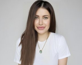 Eine Person mit langen, braunen Haaren und einem weißen T-Shirt lächelt in die Kamera, trägt eine Halskette mit einem Anhänger.