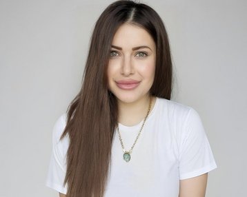 Eine Person mit langen, braunen Haaren und einem weißen T-Shirt lächelt in die Kamera, trägt eine Halskette mit einem Anhänger.