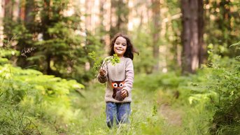 Ein Mädchen mit langen, braunen Haaren trägt einen Pullover mit einem Eulenmotiv und hält einen Zweig in der Hand, während es fröhlich einen Waldweg entlangläuft.