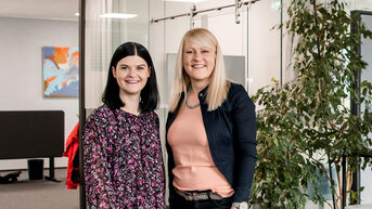Zwei Frauen stehen in einem modernen Büro mit Pflanzen und Schreibtischen. Die Frau links trägt ein blumiges Kleid, die Frau rechts eine Bluse und eine Jacke. Im Hintergrund sind weitere Büromöbel sichtbar.