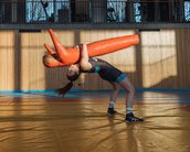Eine Person in Sportkleidung führt eine Wurftechnik mit einem Trainingsdummy in einer Sporthalle aus. Der Dummy ist orange und wird über den Kopf der Person gehalten.