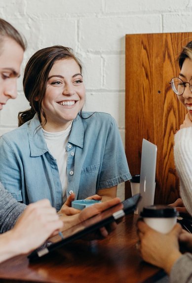 Vier Personen sitzen an einem Tisch in einem Café, lächeln und unterhalten sich. Eine Person nutzt ein Tablet, während die anderen Getränke in den Händen halten.