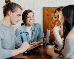 Vier Personen sitzen an einem Tisch in einem Café, lächeln und unterhalten sich. Eine Person nutzt ein Tablet, während die anderen Getränke in den Händen halten.
