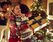 Eine Frau umarmt einen Mann, während sie ein Geschenk in der Hand hält. Im Hintergrund ist ein festlich geschmückter Weihnachtsbaum und weitere Personen, die miteinander interagieren.