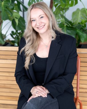 Eine lächelnde Frau mit langen, blonden Haaren sitzt in einem schwarzen Blazer und Jeans auf einem Stuhl vor einer Holzpaneelwand und Pflanzen.