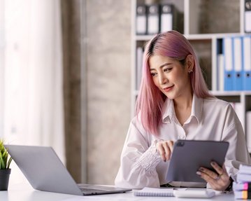 Eine Frau mit pinken Haaren sitzt an einem Schreibtisch, nutzt ein Tablet und schaut auf einen Laptop. Im Hintergrund sind Aktenordner und eine kleine Pflanze sichtbar.