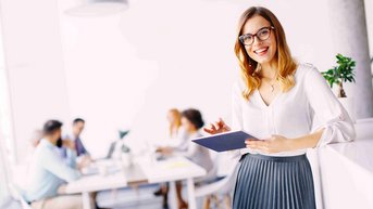Eine lächelnde Frau mit Brille hält ein Tablet in einem modernen Büro, während im Hintergrund eine Gruppe von Menschen an einem Tisch arbeitet.