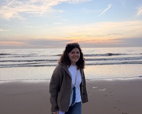 Eine Person mit lockigem Haar trägt einen grauen Pullover und Jeans und geht am Strand entlang, während die Wellen sanft an den Ufer schlagen.
