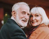Ein älteres Paar lächelt sich an, während sie sich umdrehen. Der Mann hat graue Haare und einen Bart, die Frau hat schulterlanges, helles Haar. Beide tragen warme Kleidung und stehen in einer gemütlichen Umgebung.